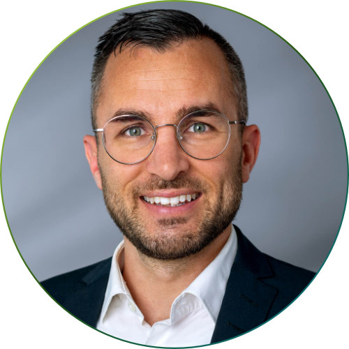 Nahaufnahme eines lächelnden Mannes mit kurzem braunem Haar und Bart, der ein hellblaues Hemd trägt, vor unscharf grünem Außenhintergrund.