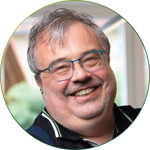 Mann mit kurzem braunem Haar und hellem Hemd trägt dunkelblauen karierten Blazer und lächelt in die Kamera.