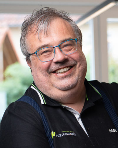 Porträt eines lächelnden Mannes mit kurzen braunen Haaren, in einem weißen Hemd und dunkelgrauem kariertem Sakko.