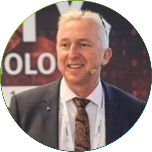 Nahaufnahme eines lächelnden Mannes mit kurzem braunem Haar und Bart, der ein hellblaues Hemd trägt, vor unscharf grünem Außenhintergrund.