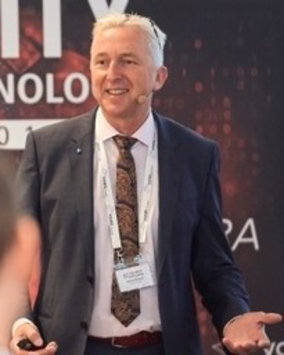 Porträt eines lächelnden Mannes mit kurzen braunen Haaren, in einem weißen Hemd und dunkelgrauem kariertem Sakko.