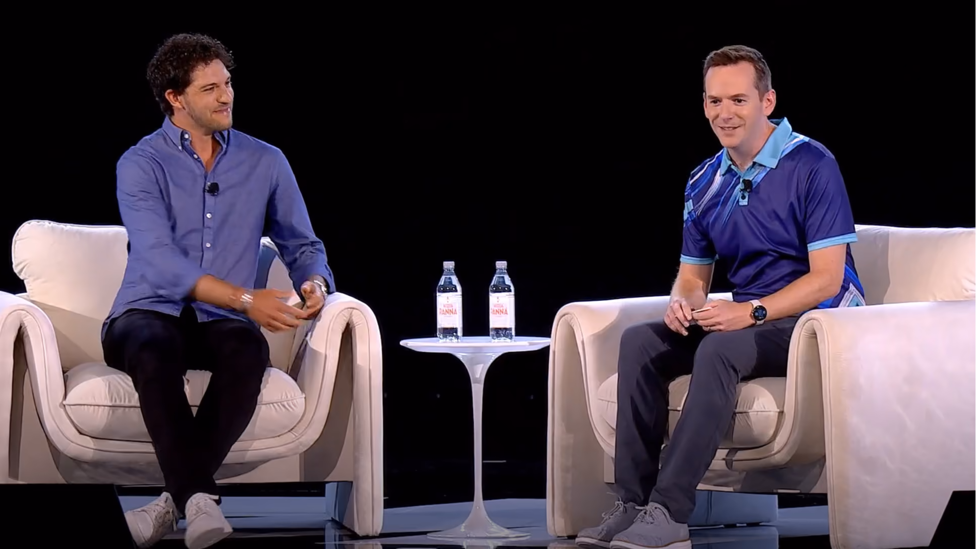 Two men seated in white armchairs on a stage with a small round table, engaged in conversation.