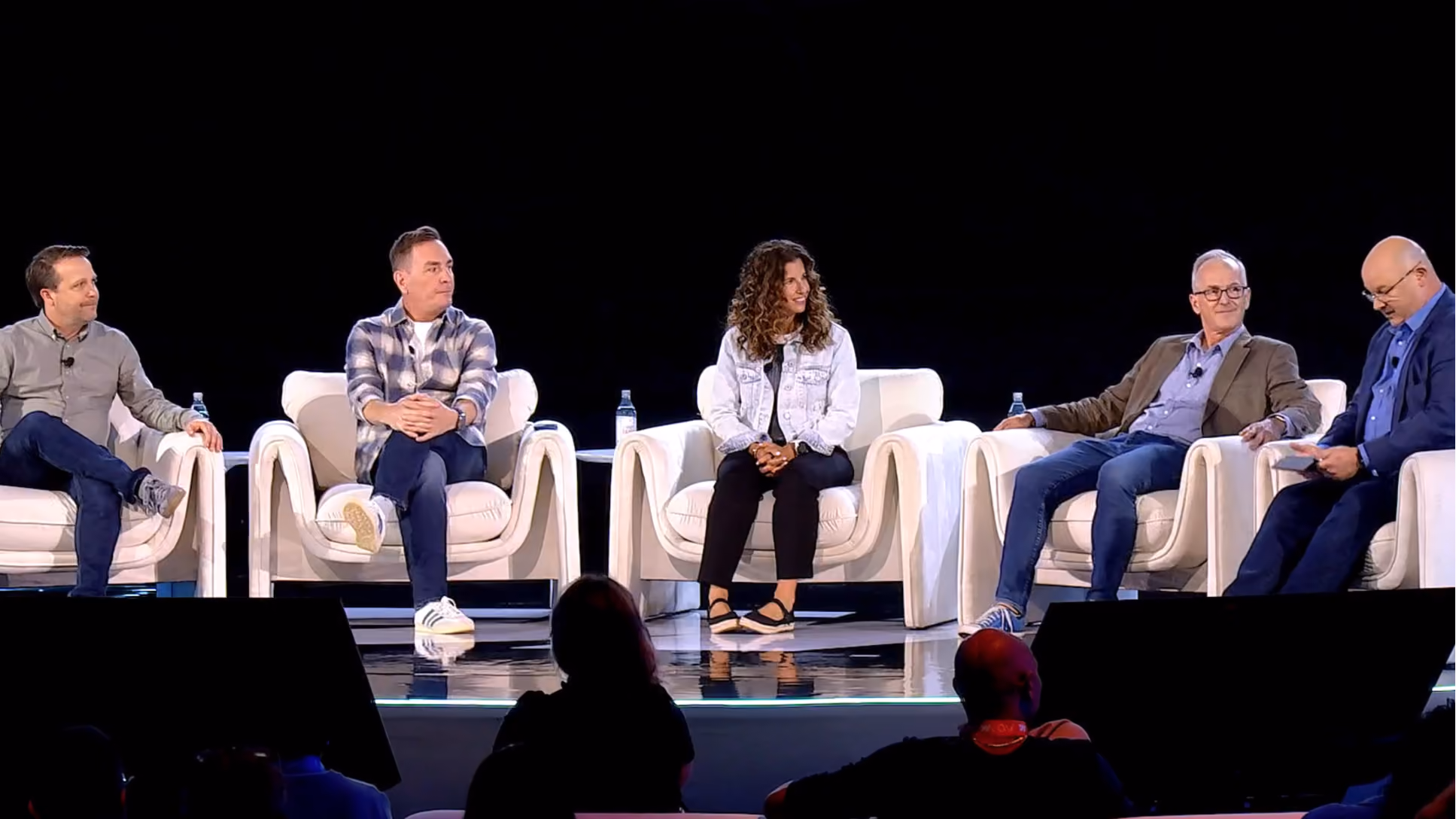 Five people sitting in white armchairs on a stage with a black background, engaging in a panel discussion.