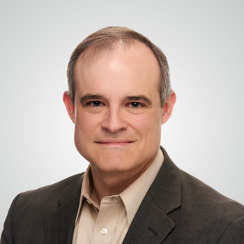 Portrait of Michael Daniel wearing dark brown suit over beige dress shirt in front of pale gray backdrop