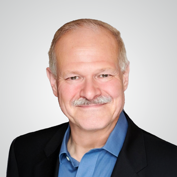 Portrait of Jim Richberg wearing black suit and kelly blue dress shirt in front of pale gray backdrop