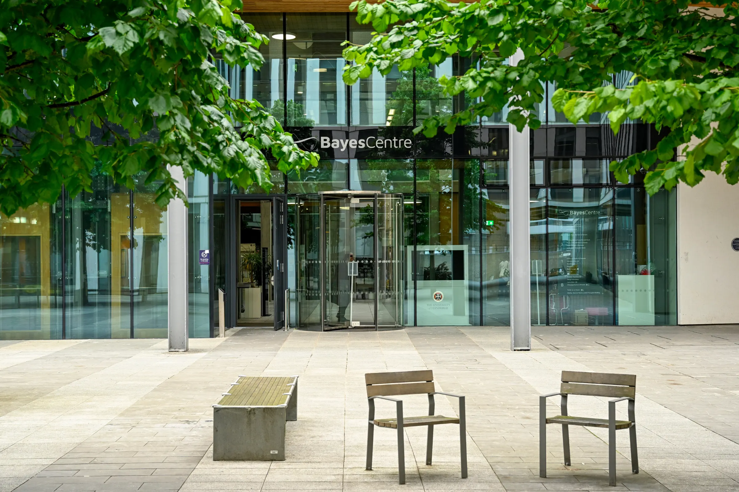 Exterior view of the Bayes Centre building entrance with glass doors, benches, and two chairs under green tree branches.