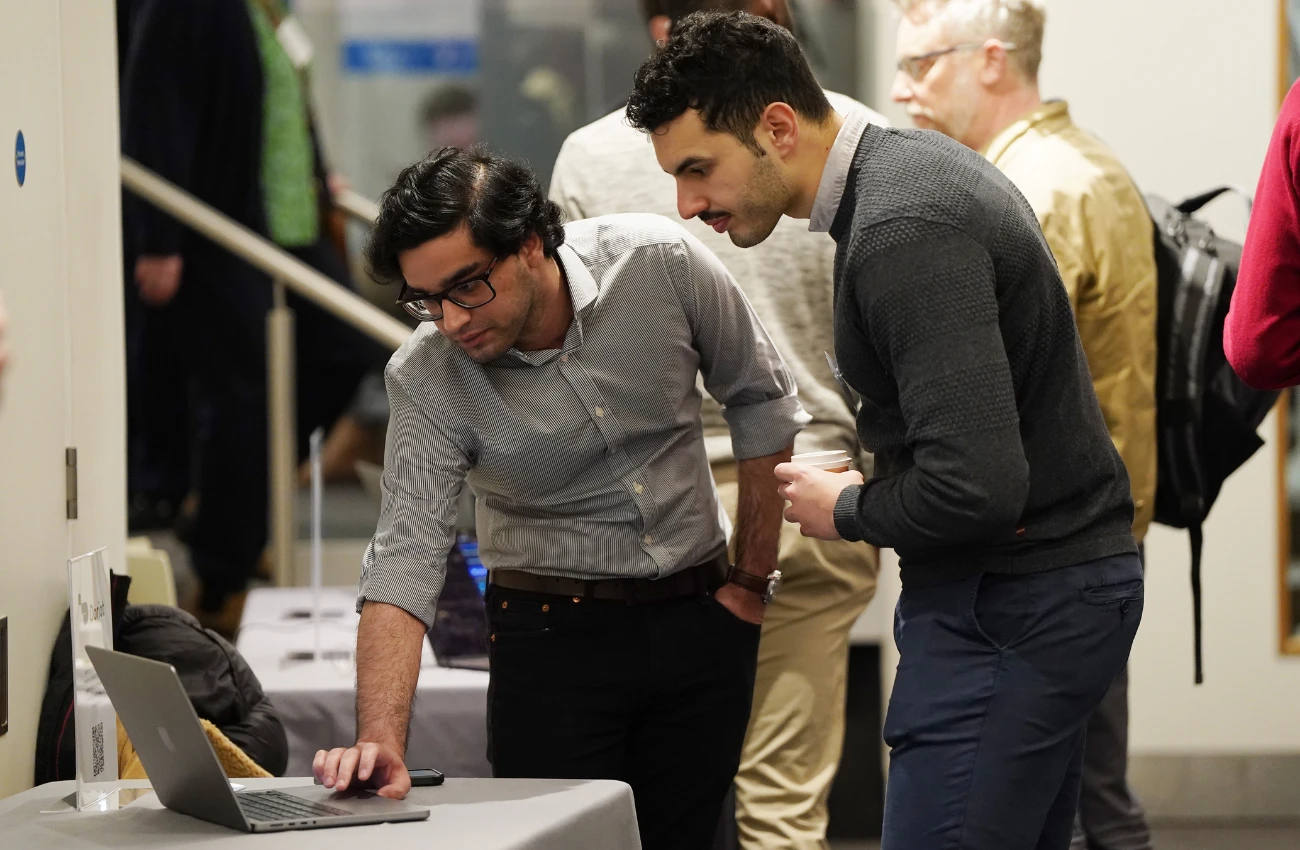 Two men looking at a laptop on a table at an indoor event, with other attendees in the background.