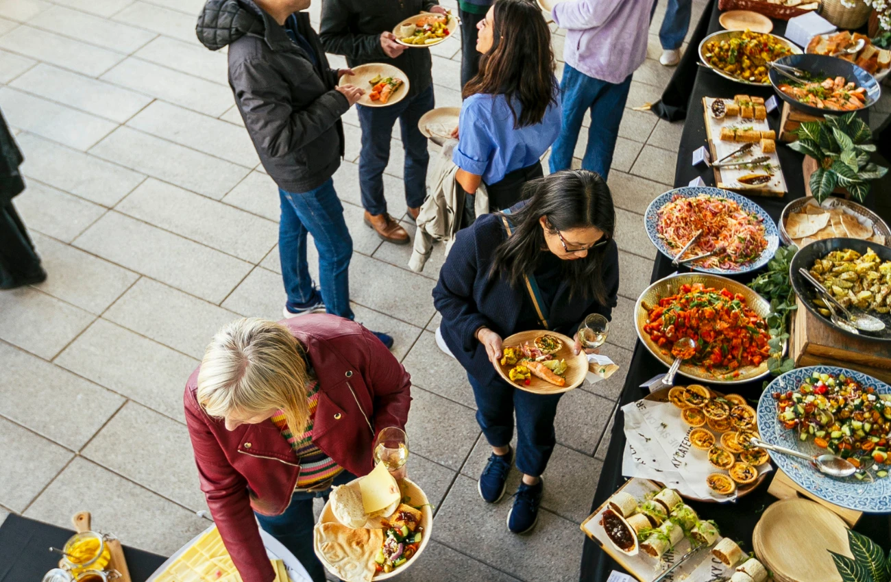 Group of people serving themselves from a buffet table with various dishes including salads, rolls, and cooked vegetables.