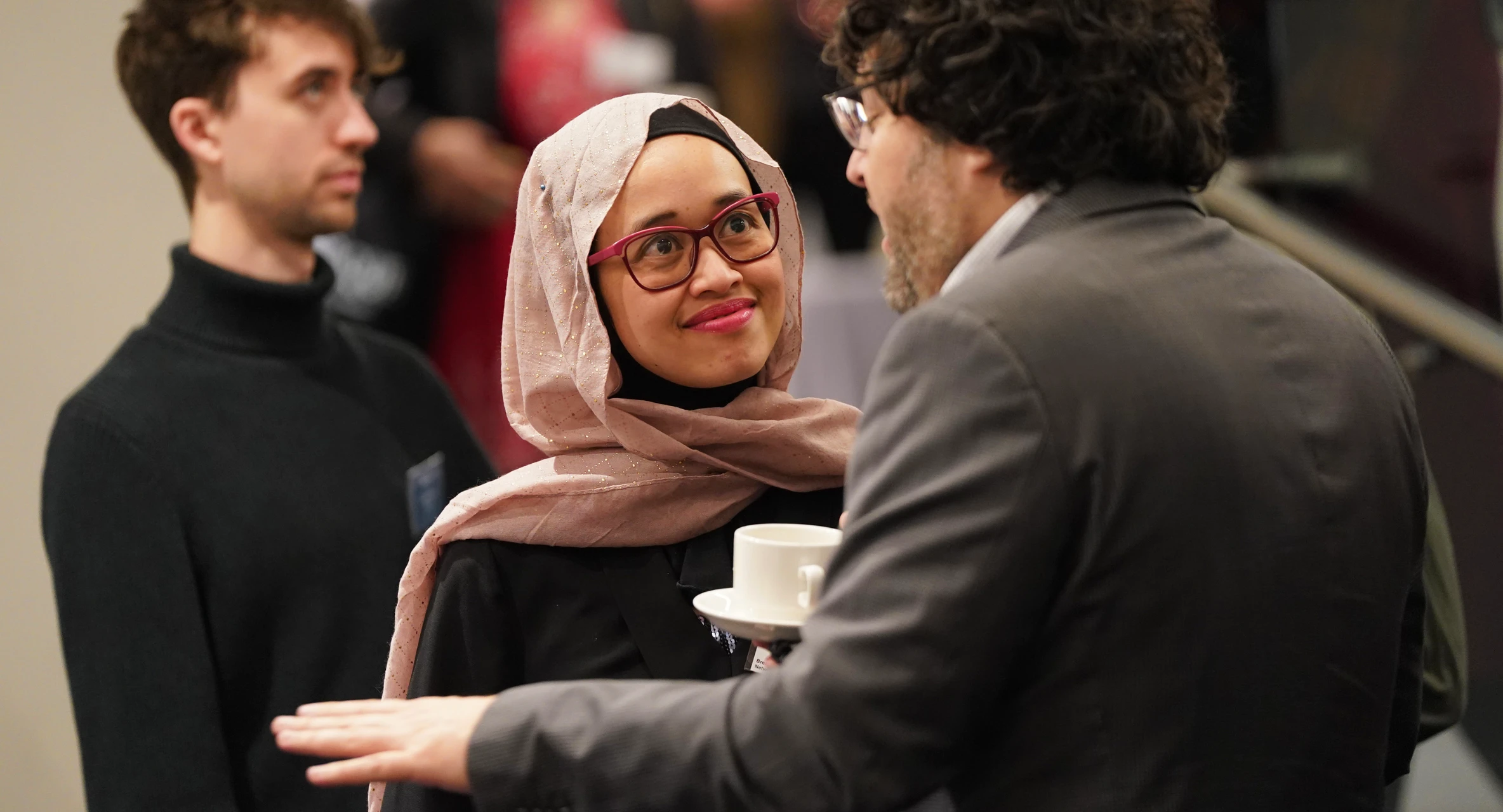 Woman wearing a pink hijab and glasses attentively listening to a man in a grey suit holding a white cup and saucer.