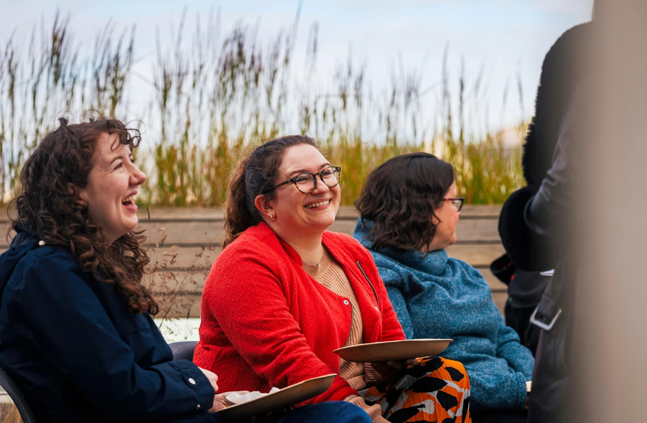 Three women sitting outdoors on benches, two smiling and holding plates, with tall grass in the background.