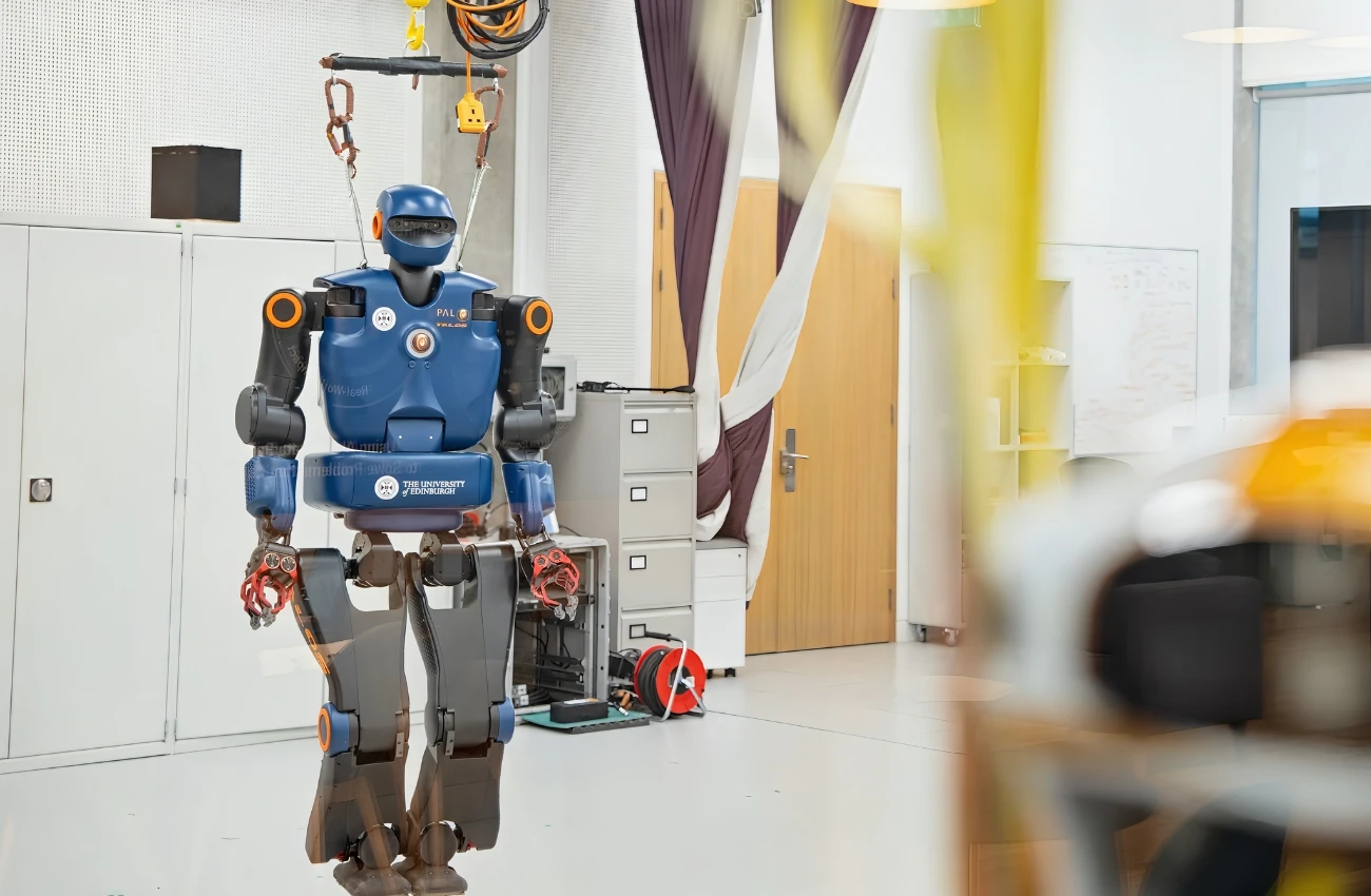 Blue humanoid robot with orange accents suspended by cables in a modern laboratory room with cabinets and a wooden door.