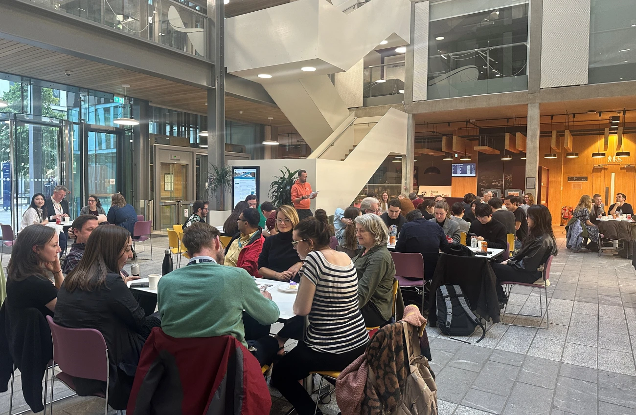 Large group of people sitting at tables in a modern indoor public space with glass walls and a white staircase.
