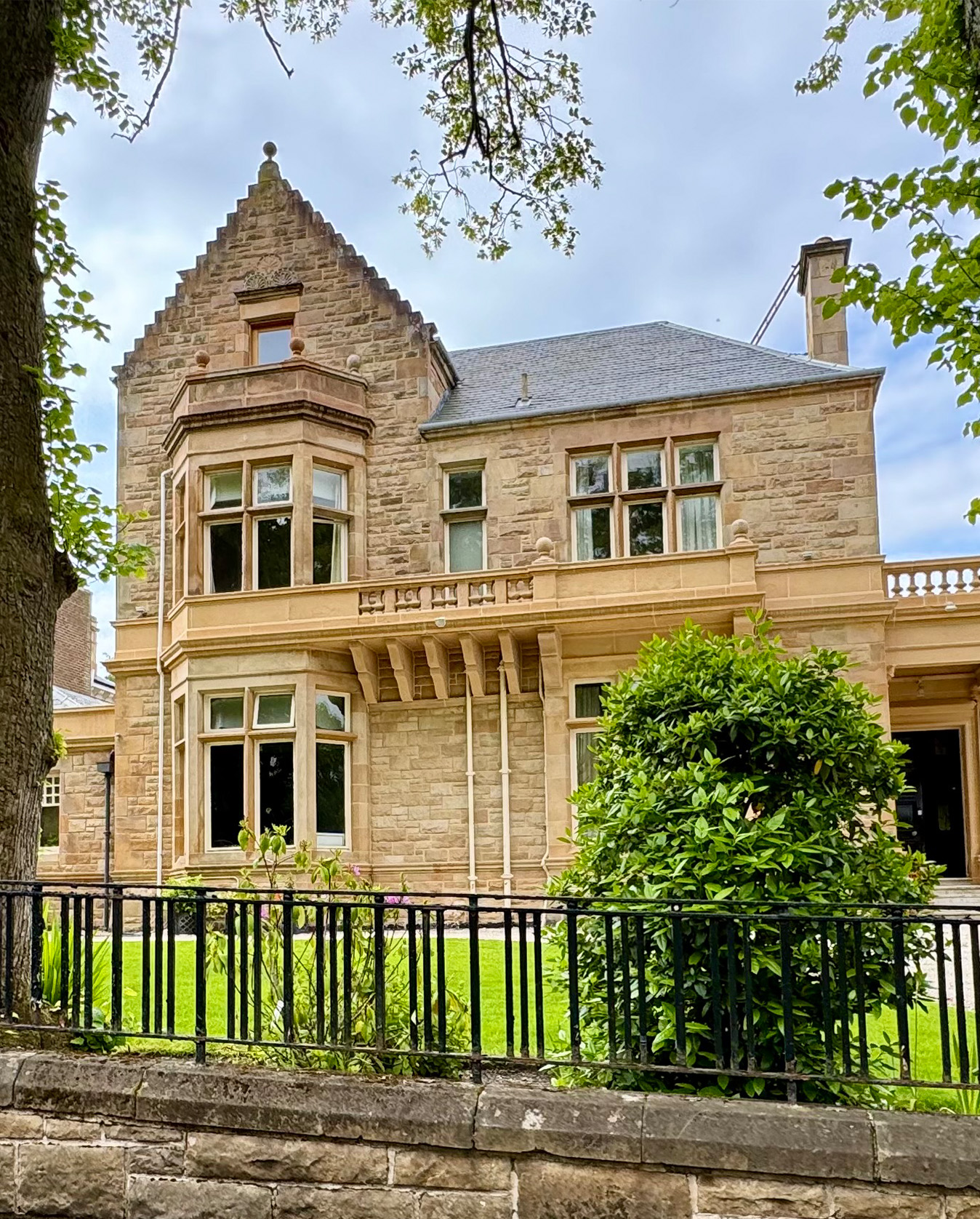 Stone house with tall windows, bay window, green lawn, bushes, and iron fence in front.