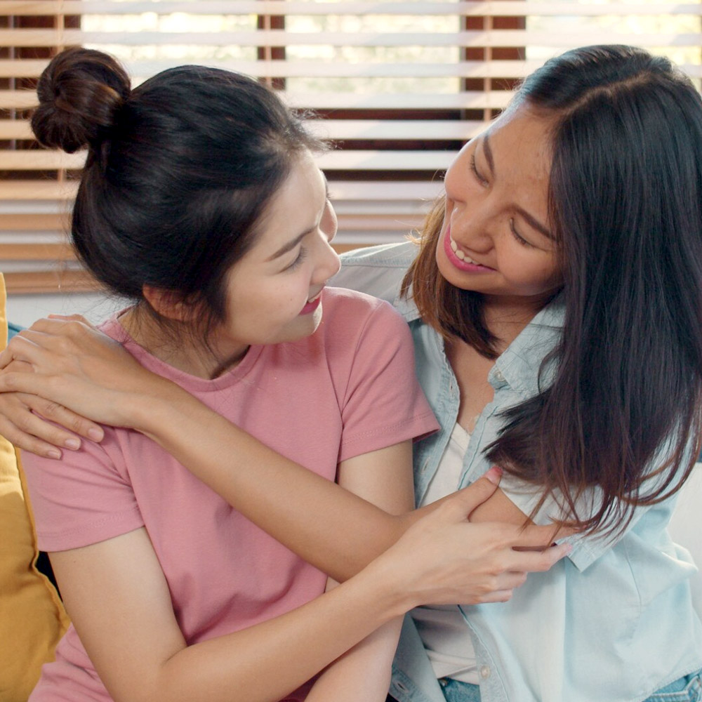 Two Asian women sharing a warm, supportive embrace while seated together at home.