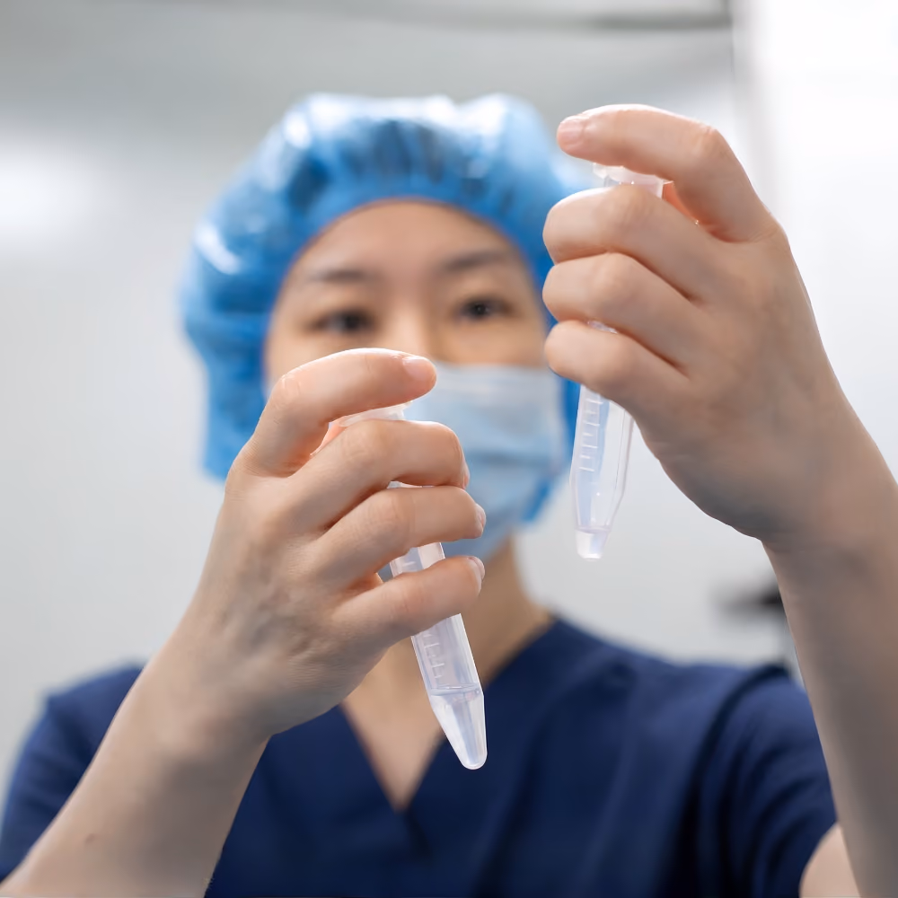 Laboratory professional in navy scrubs and protective equipment inspecting two sample tubes in a fertility laboratory.