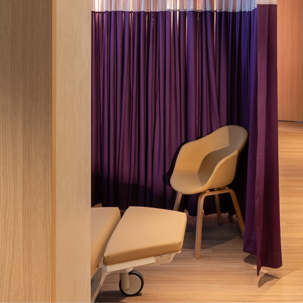 GenPrime Fertility treatment area with an adjustable medical bed, visitor chair, purple privacy curtains, and warm wood finishes.