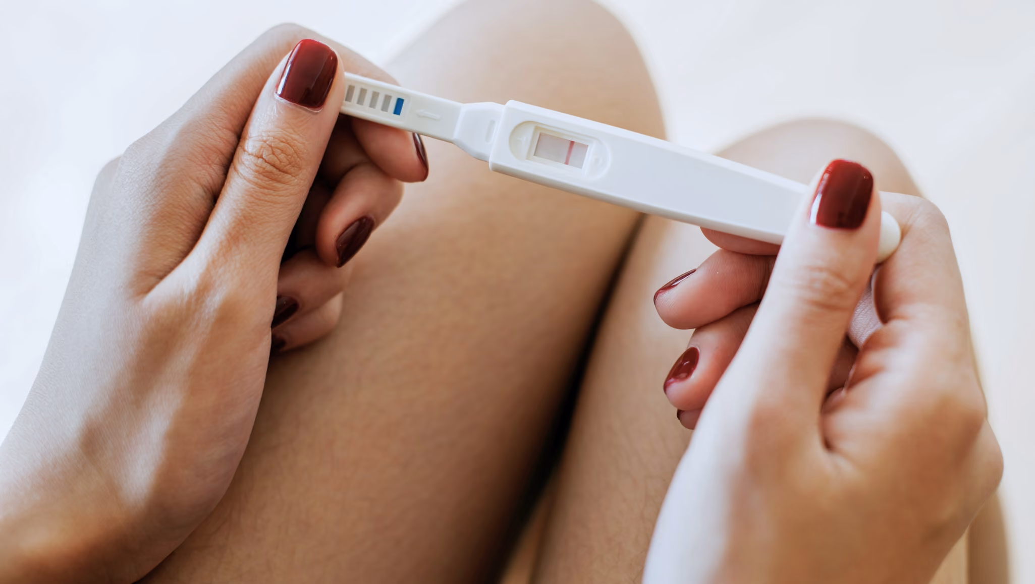 Hands holding a positive pregnancy test while seated against a bright, neutral background.