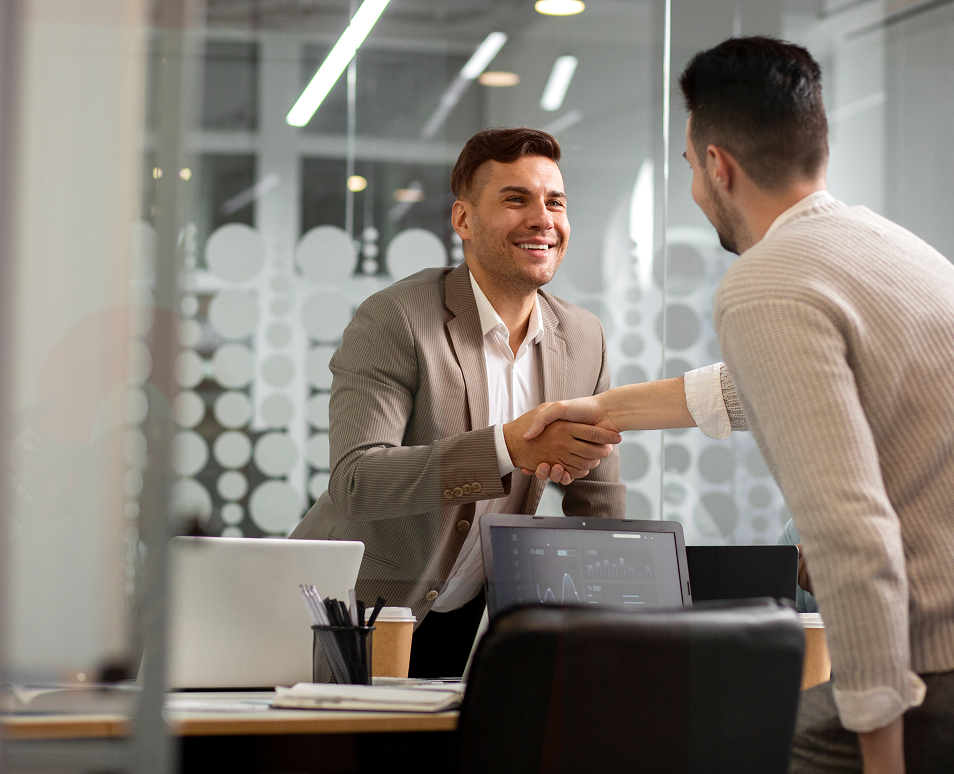 Two men shaking hands across an office table with laptops and coffee cups, smiling during a business meeting.