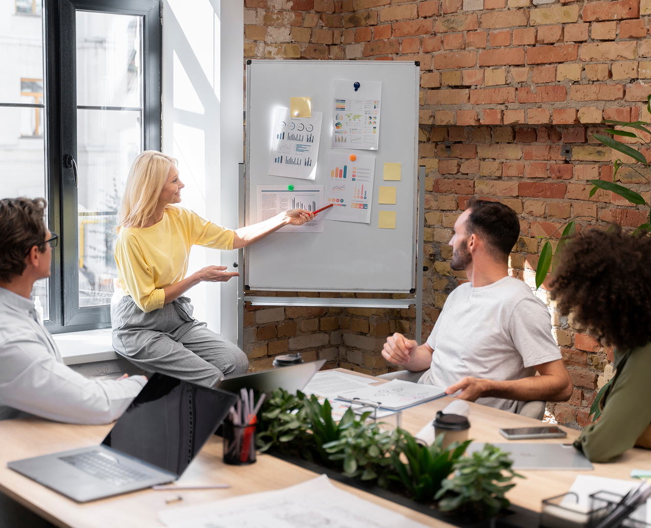 A woman in a yellow shirt presenting charts on a whiteboard to three seated colleagues in a bright office with brick walls.