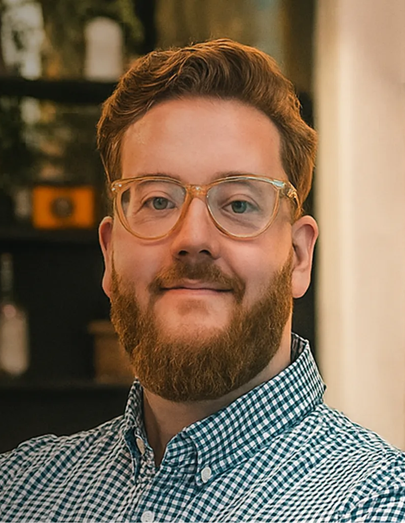 Man with a red beard and glasses wearing a blue checkered shirt, smiling slightly.