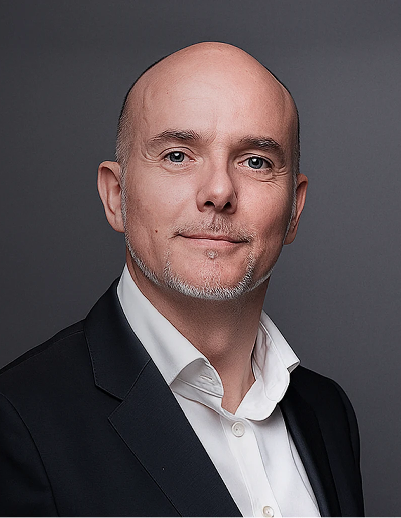 Portrait of a middle-aged bald man with light facial hair wearing a white shirt and black suit jacket, against a gray background.