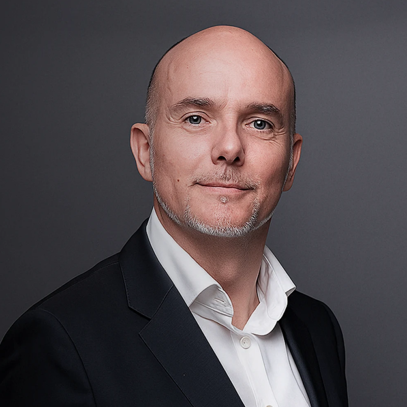 Portrait of a bald man with light facial hair wearing a white dress shirt and black blazer, against a gray background.