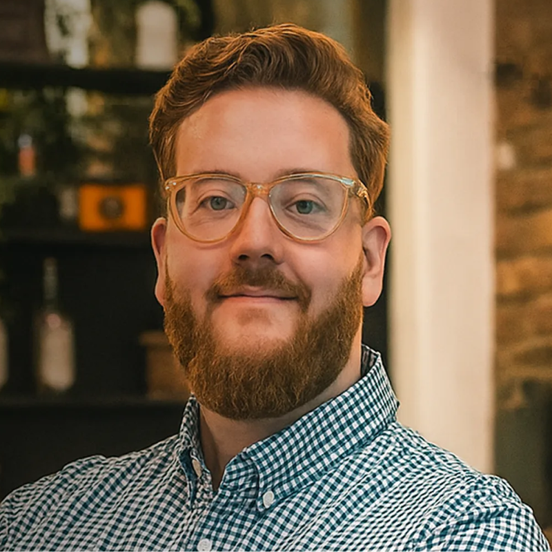 Man with a red beard and glasses wearing a blue and white checkered shirt, smiling indoors.