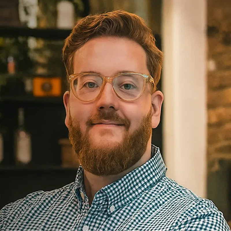 Man with a red beard and glasses wearing a blue and white checkered shirt, smiling indoors.