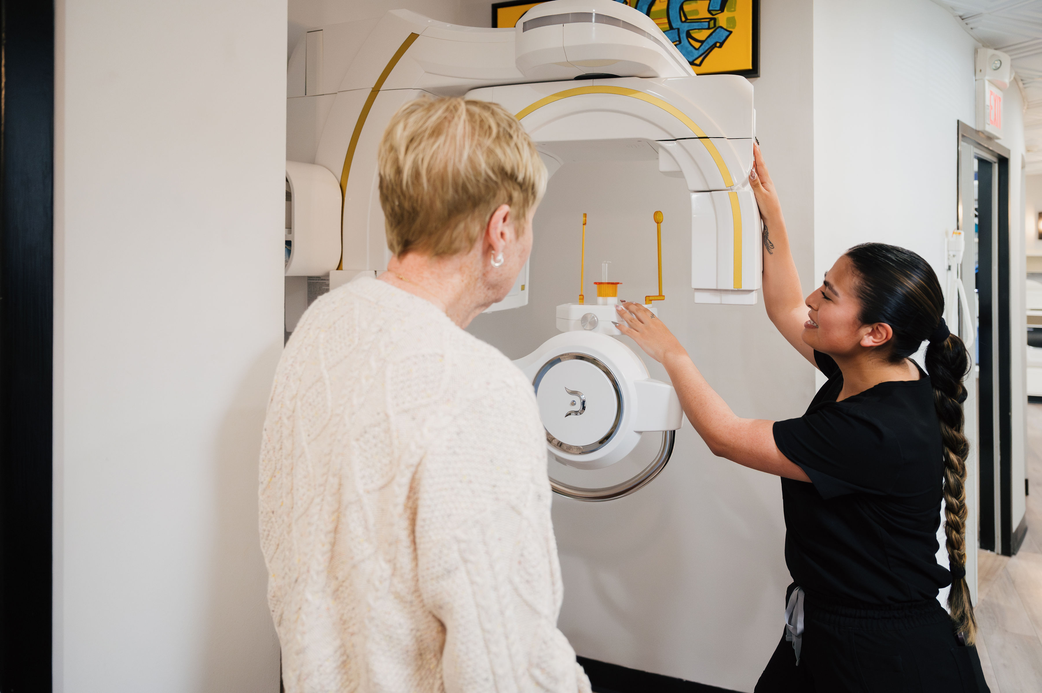 A healthcare worker, possibly a dentist, explains the operation of a medical imaging machine to an older woman in a clinical setting.