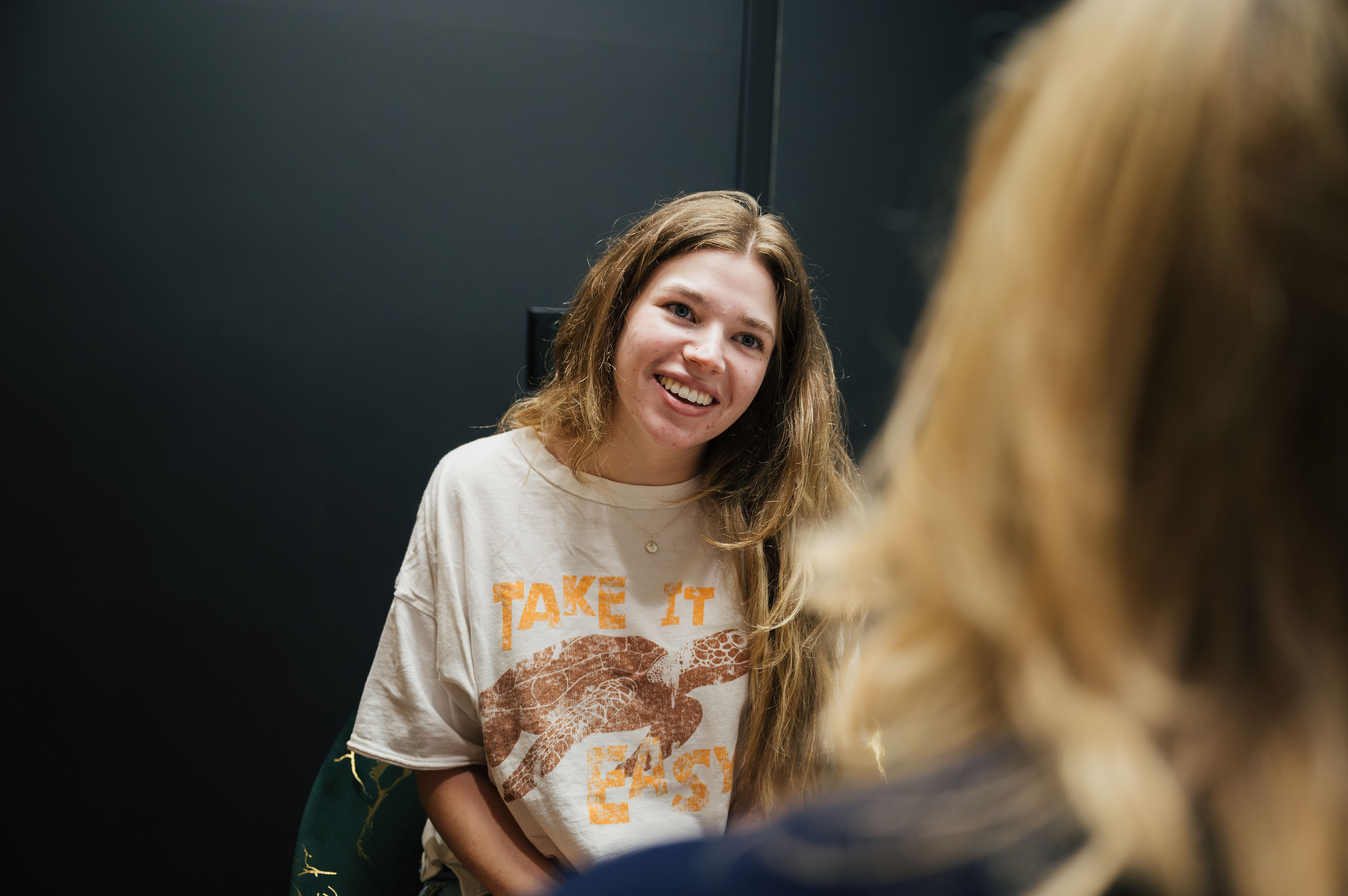A young woman with long hair, wearing a "Take it Easy" t-shirt, smiles while talking to a dentist in an indoor setting.