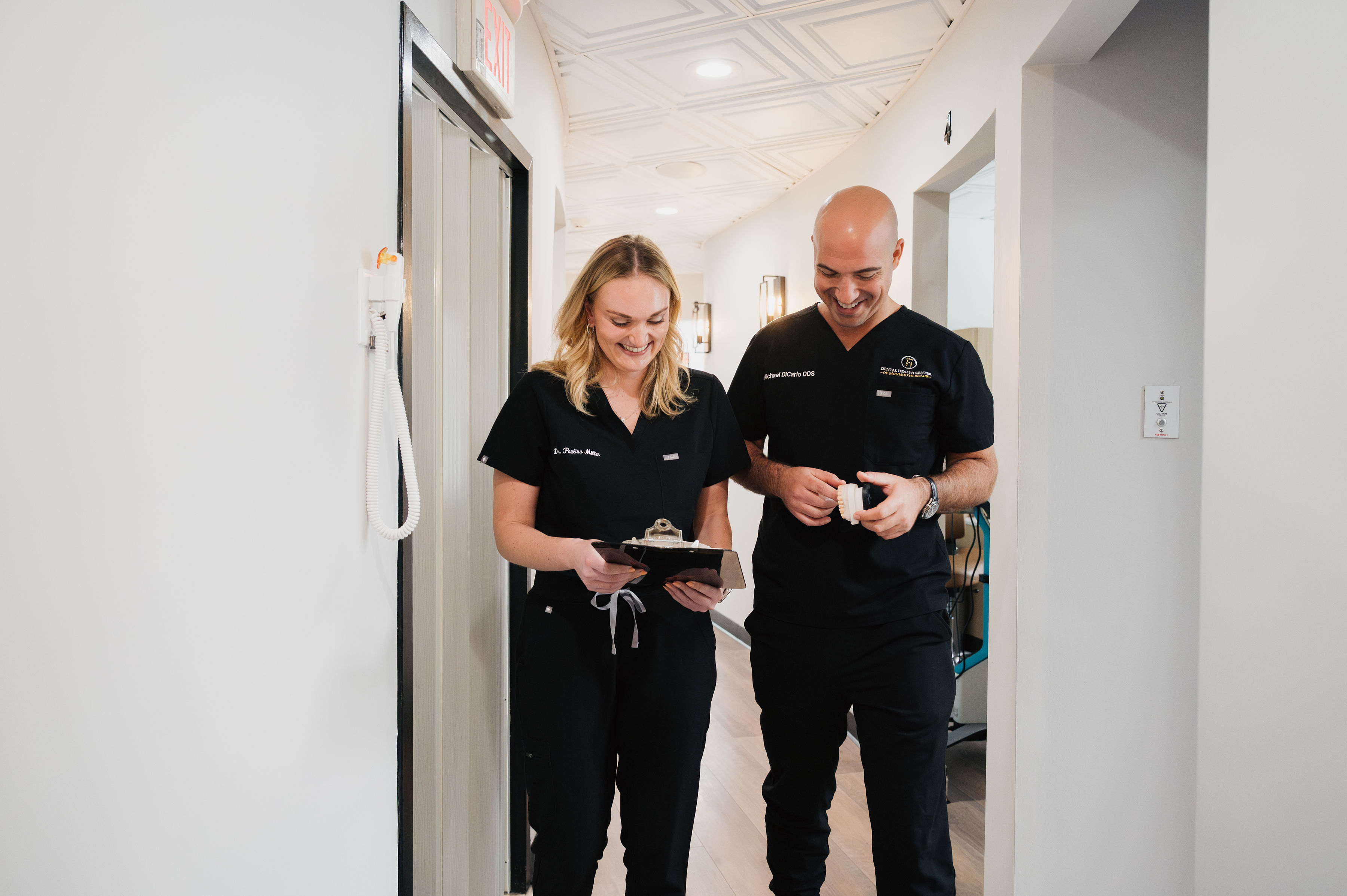 Two medical professionals, including a dentist in black scrubs, walk down a hallway, smiling as they look at a clipboard and a cup.