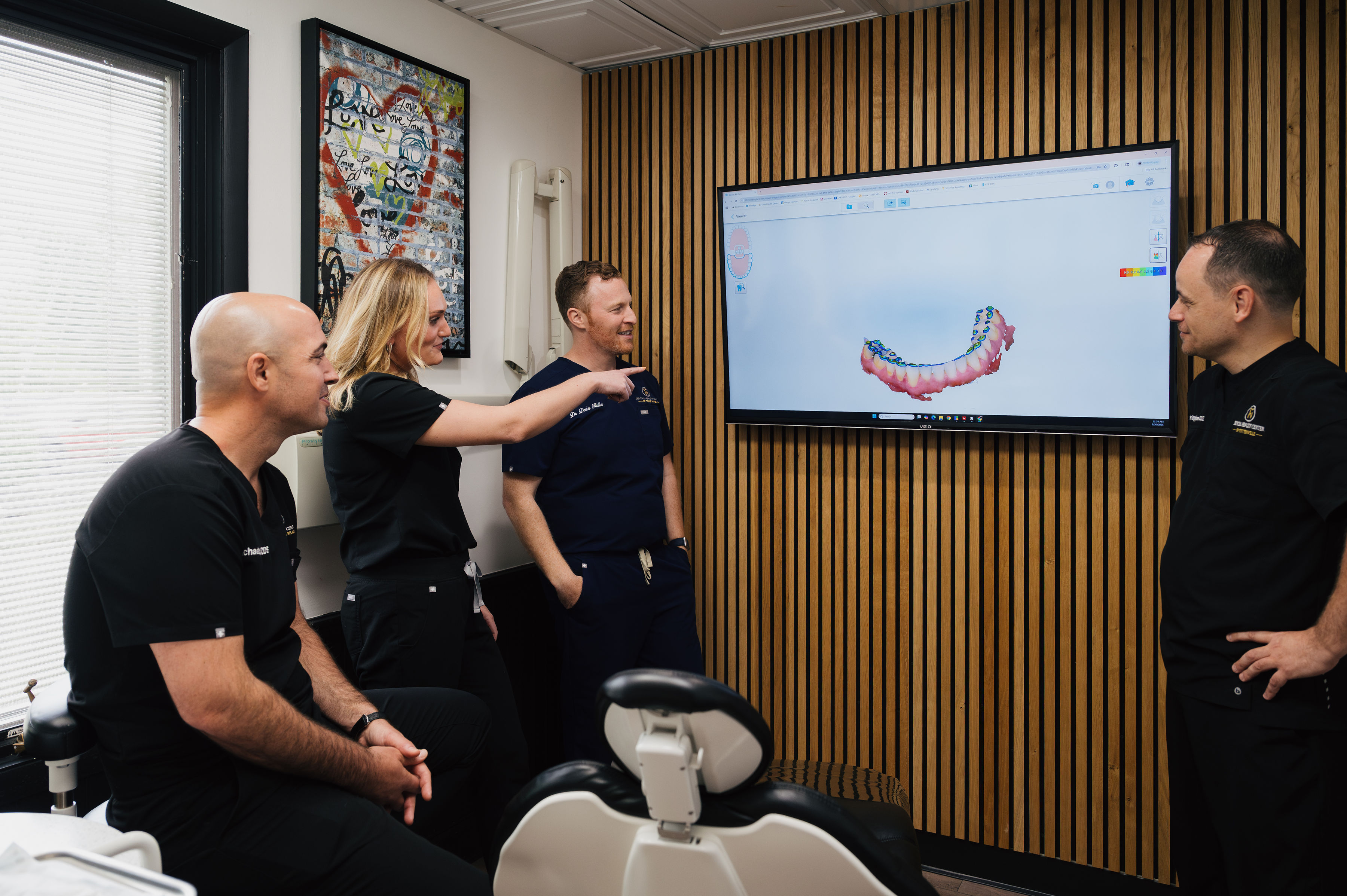 Four dentists in scrubs discuss a 3D dental model displayed on a large screen in a modern office with wood panel walls.
