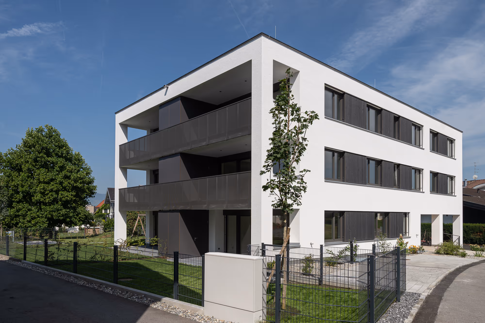 Modern white three-story building with dark balcony railings and windows under clear blue sky.