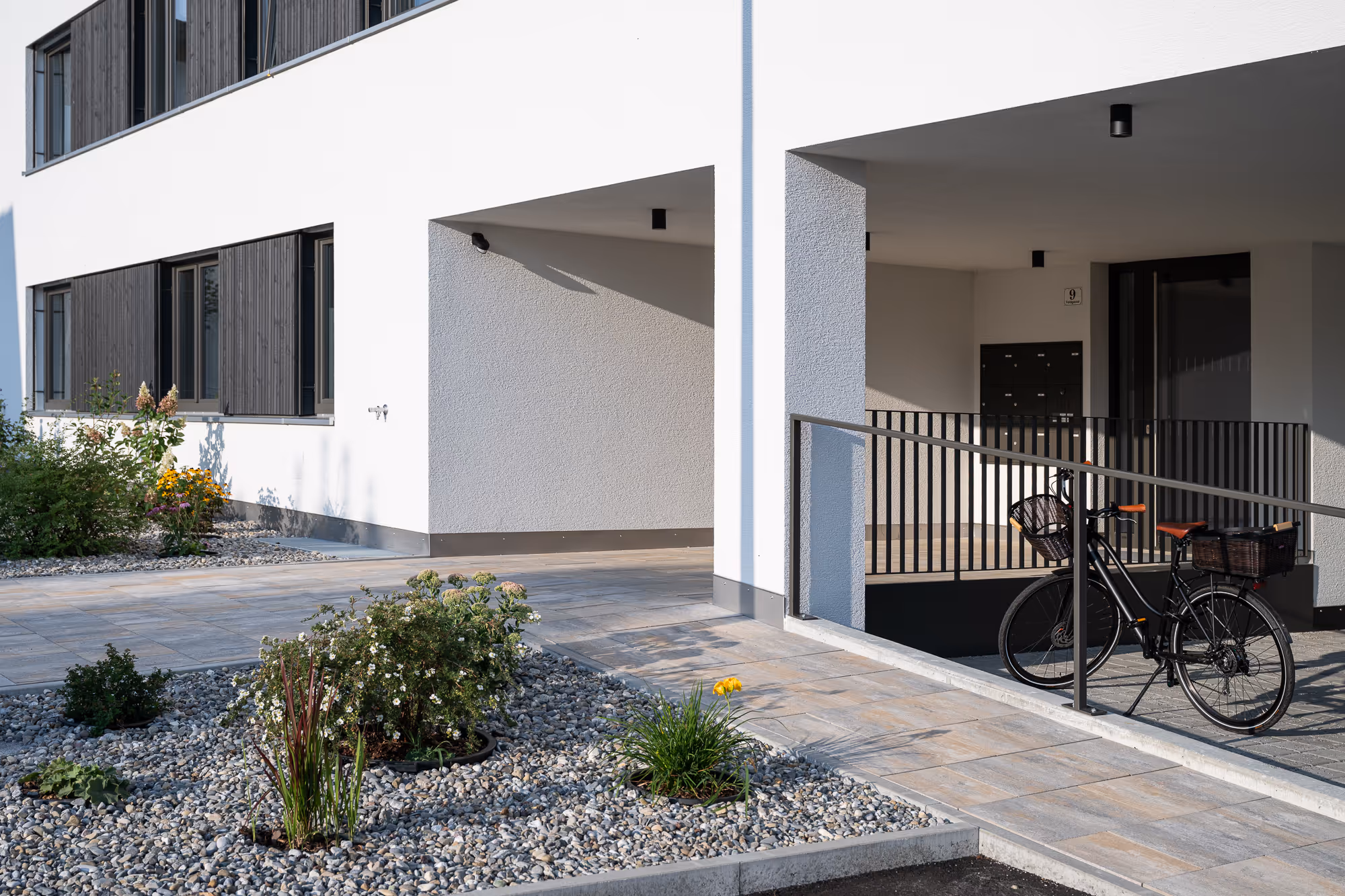 Modern white building entrance with plants, stone pavement, and bicycle with baskets parked by railing.