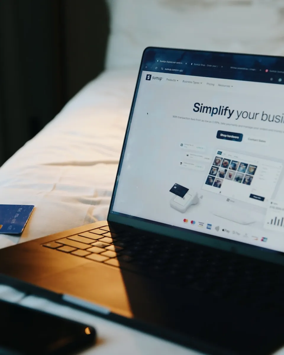A laptop displaying a webpage titled "Simplify your business" on a bed with a white sheet. A credit card is partially visible on the left side.