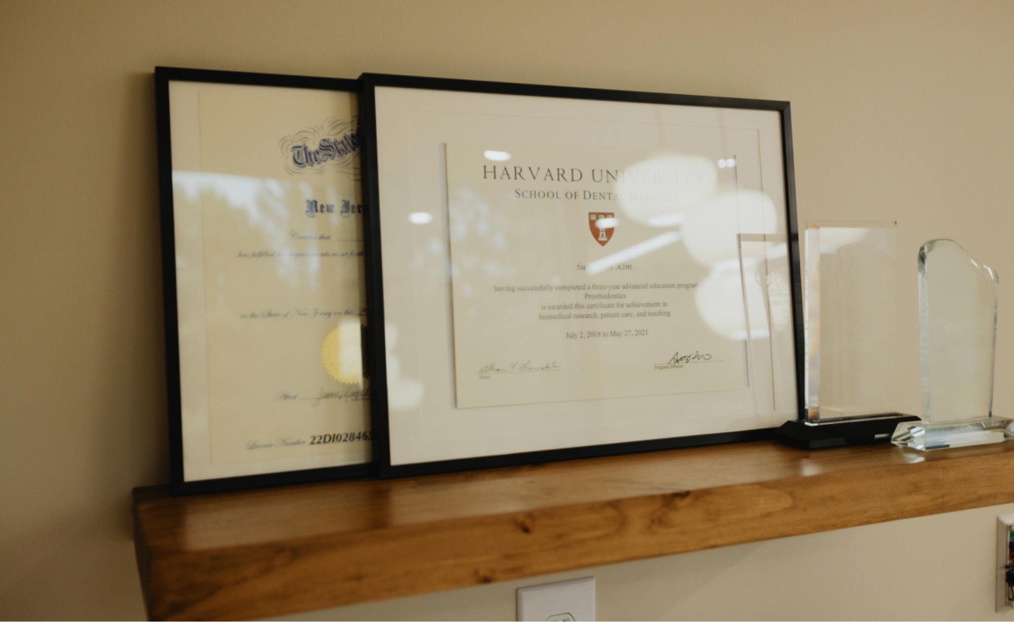 Two framed certificates, including a Harvard University School of Dental Medicine diploma, displayed on a wooden shelf next to clear glass awards at Montvale Prosthodontics.