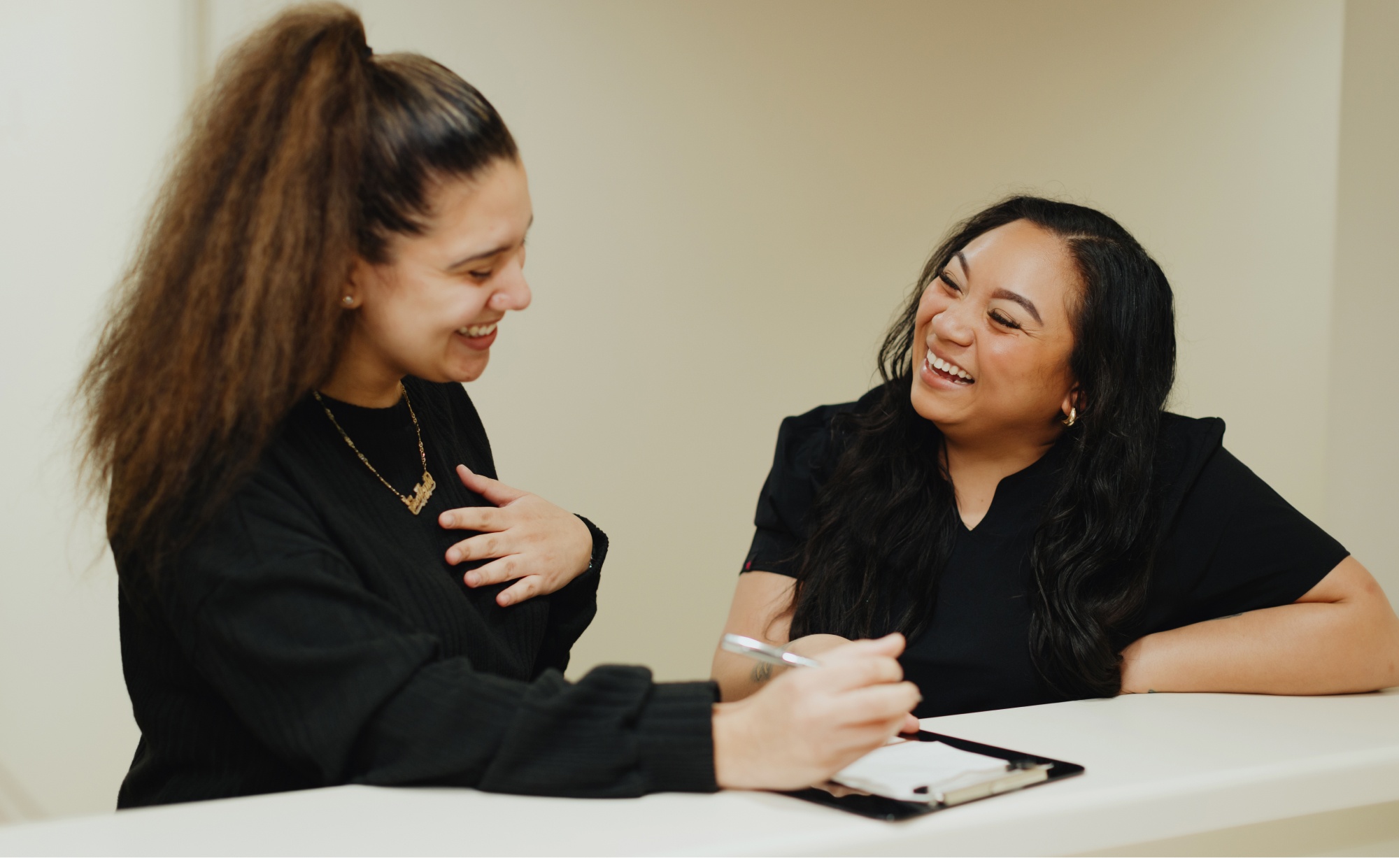 Two women are smiling and talking at a counter. One, perhaps a dentist, is holding a pen and clipboard. Both are wearing black tops.