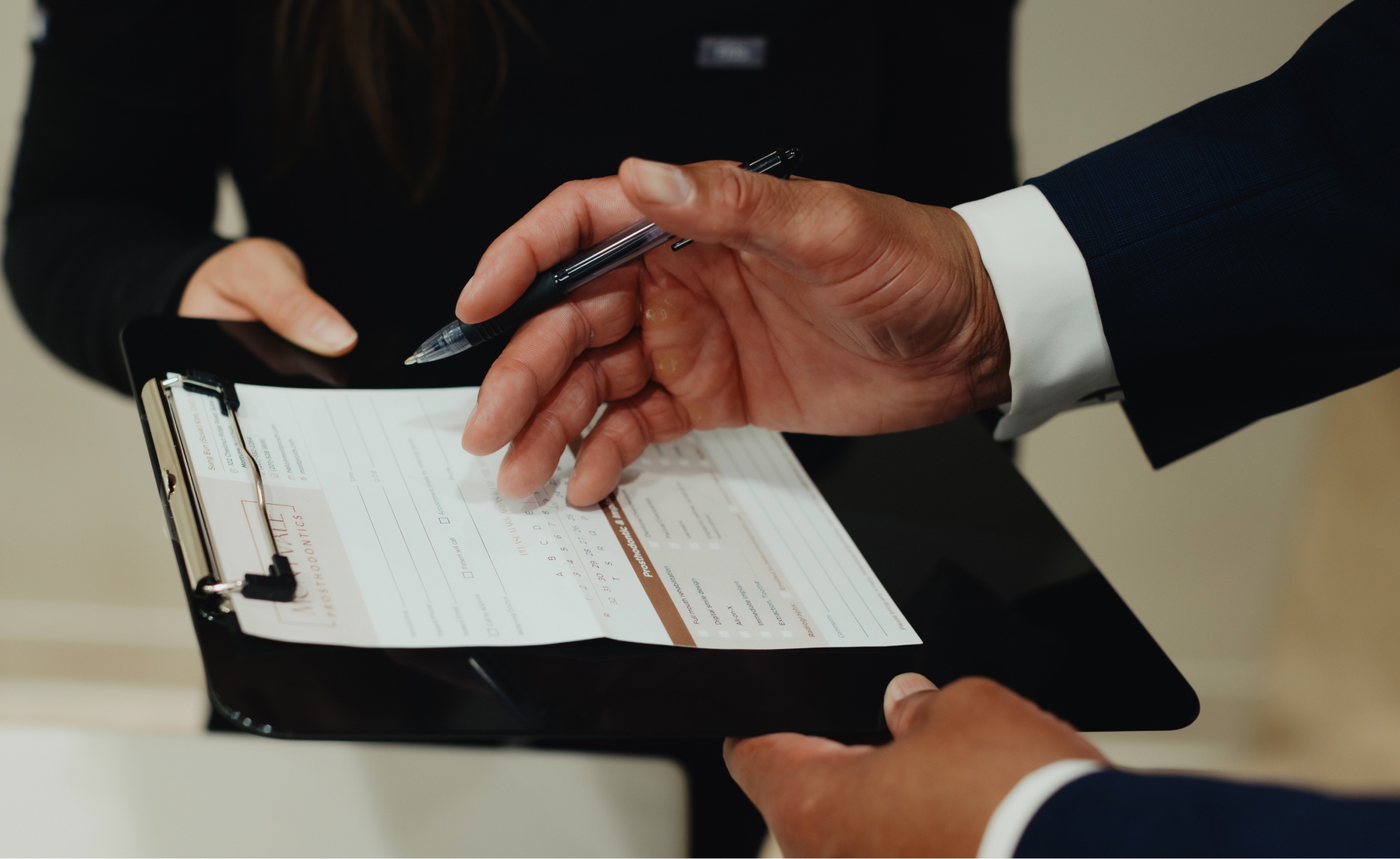 A close-up of two people holding a clipboard with a document on it. One person, perhaps a dentist, is holding a pen and gesturing towards the document.