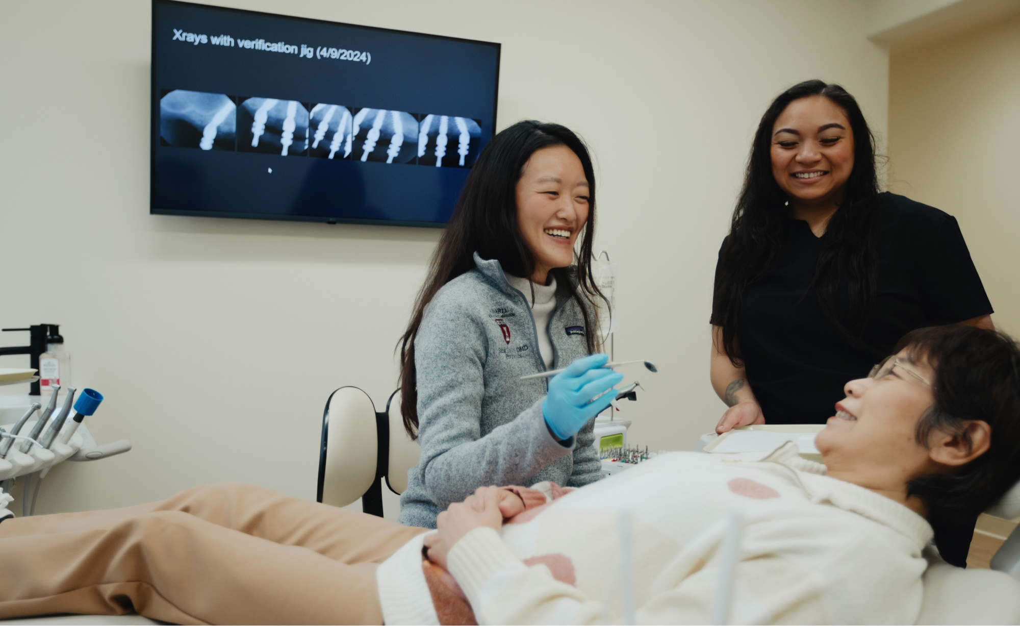 A dentist and dental assistant attend to a patient lying on a dental chair, with X-rays displayed on a monitor in the background revealing detailed dental implants.