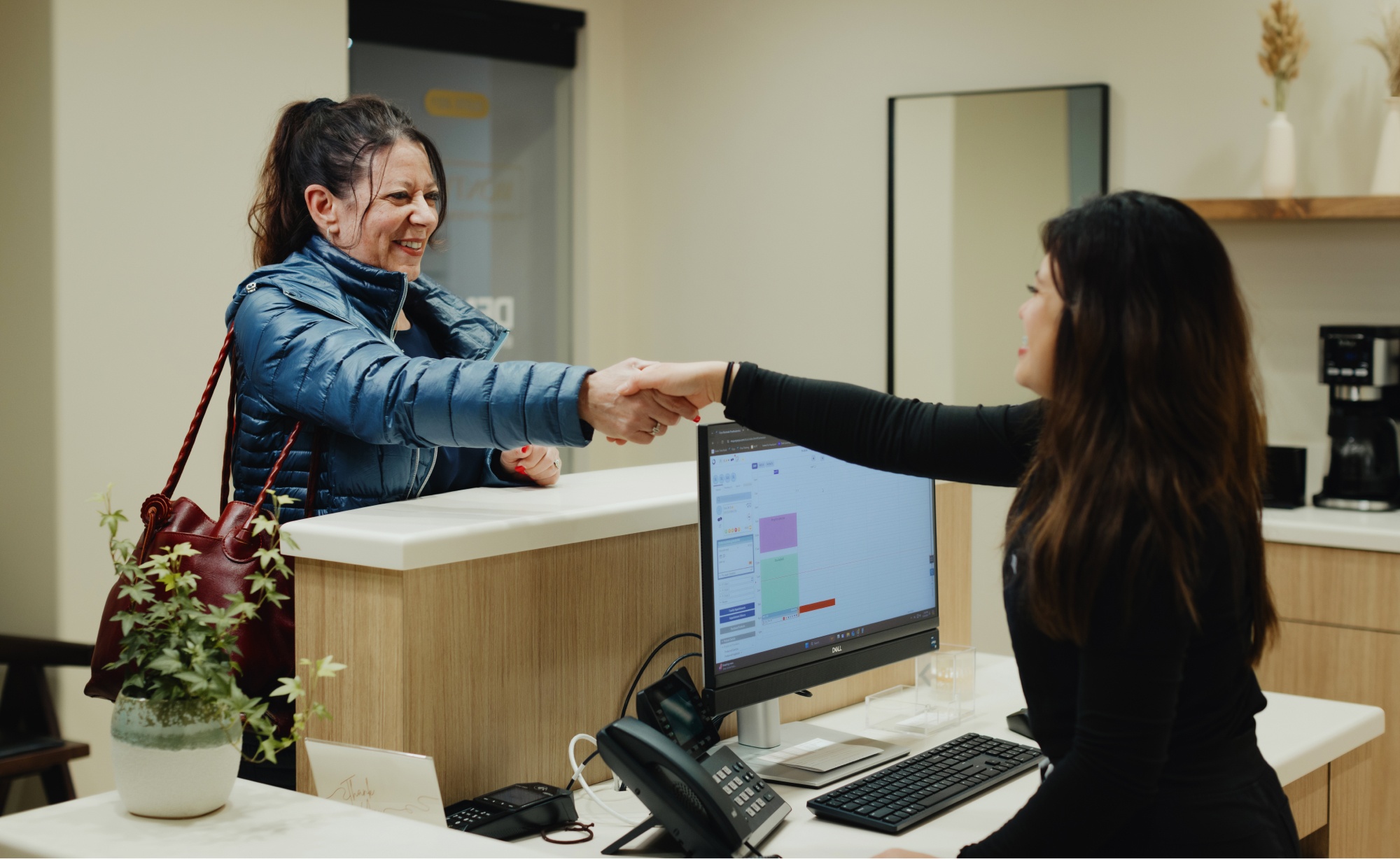 A woman in a blue jacket shakes hands with a receptionist across the counter in a dentist's office. A computer, phone, and potted plant are visible on the desk.