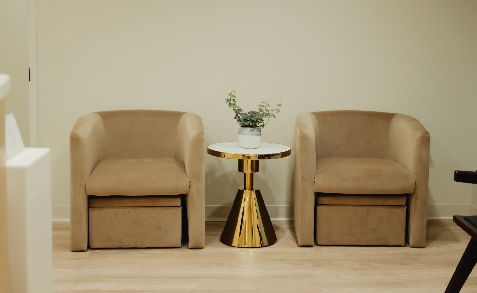 Two beige armchairs are placed with a small round table between them. The table, reminiscent of a dentist's office with its clean lines, has a gold base and a white top, holding a small potted plant. The room has light-colored walls and a wood floor.