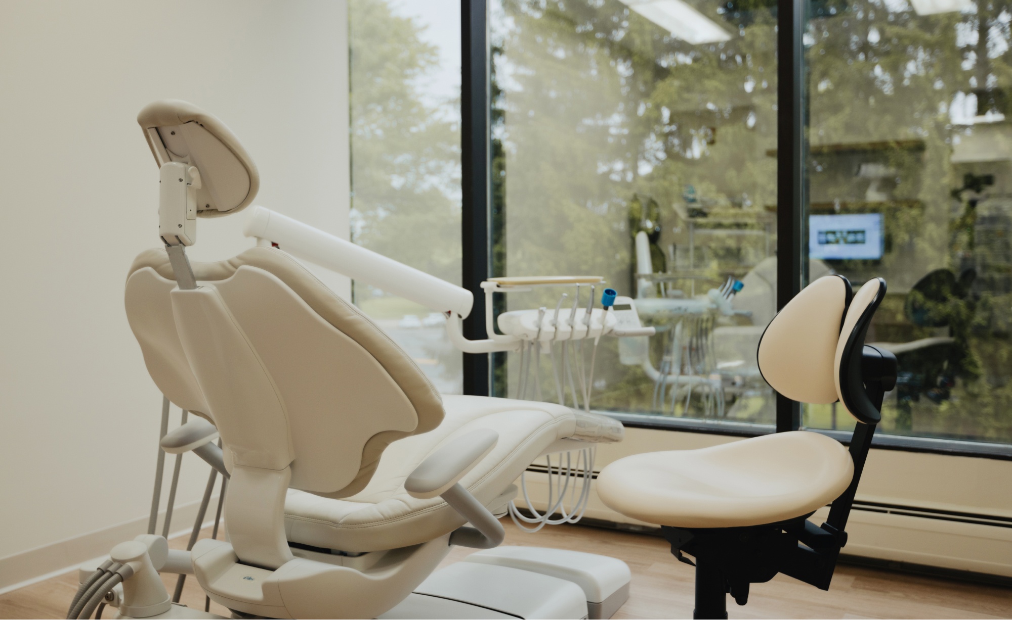 At Montvale Prosthodontics, a dental examination chair and a stool are positioned in a well-lit dental office with large windows overlooking a tree-filled outdoor area. Dental tools are carefully attached to the chair, ensuring the dentist has everything needed for optimal patient care.