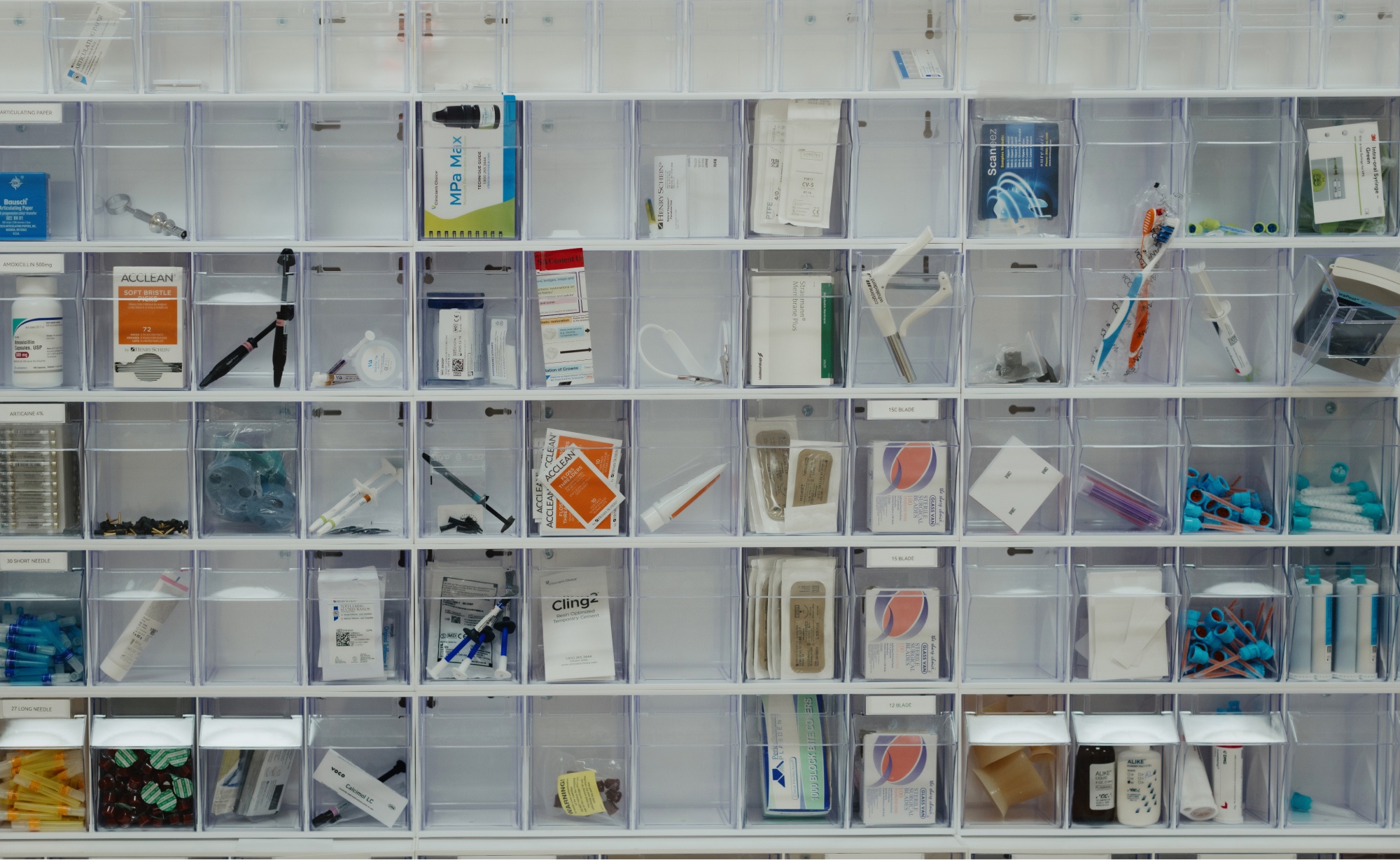 A wall-mounted organizer at Montvale Prosthodontics with clear compartments containing various medical items such as bandages, syringes, gloves, scissors, and medication boxes.