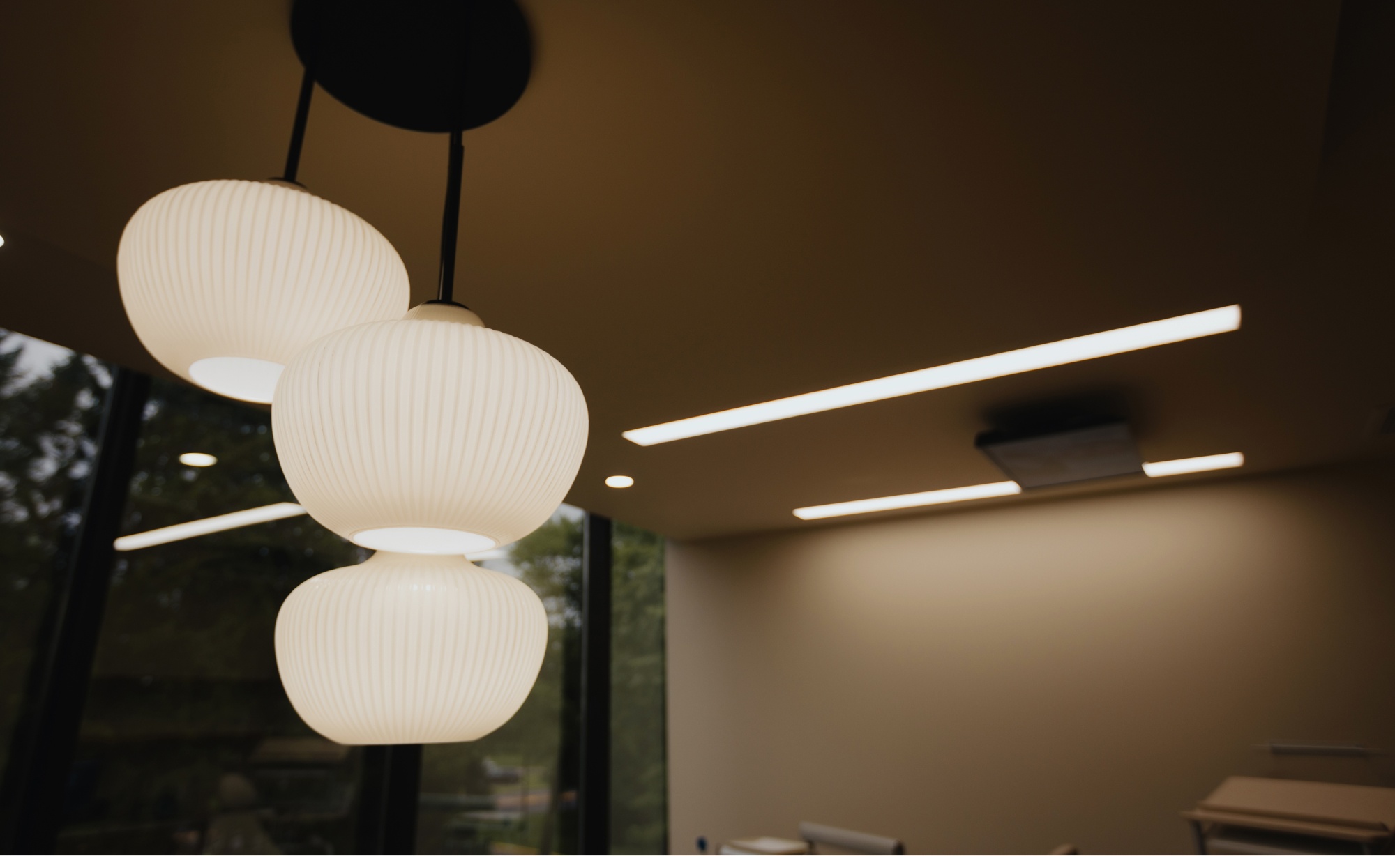 A modern ceiling light fixture with three white, ribbed lampshades hangs in a well-lit room at Montvale Prosthodontics, complete with windows and additional linear lighting.