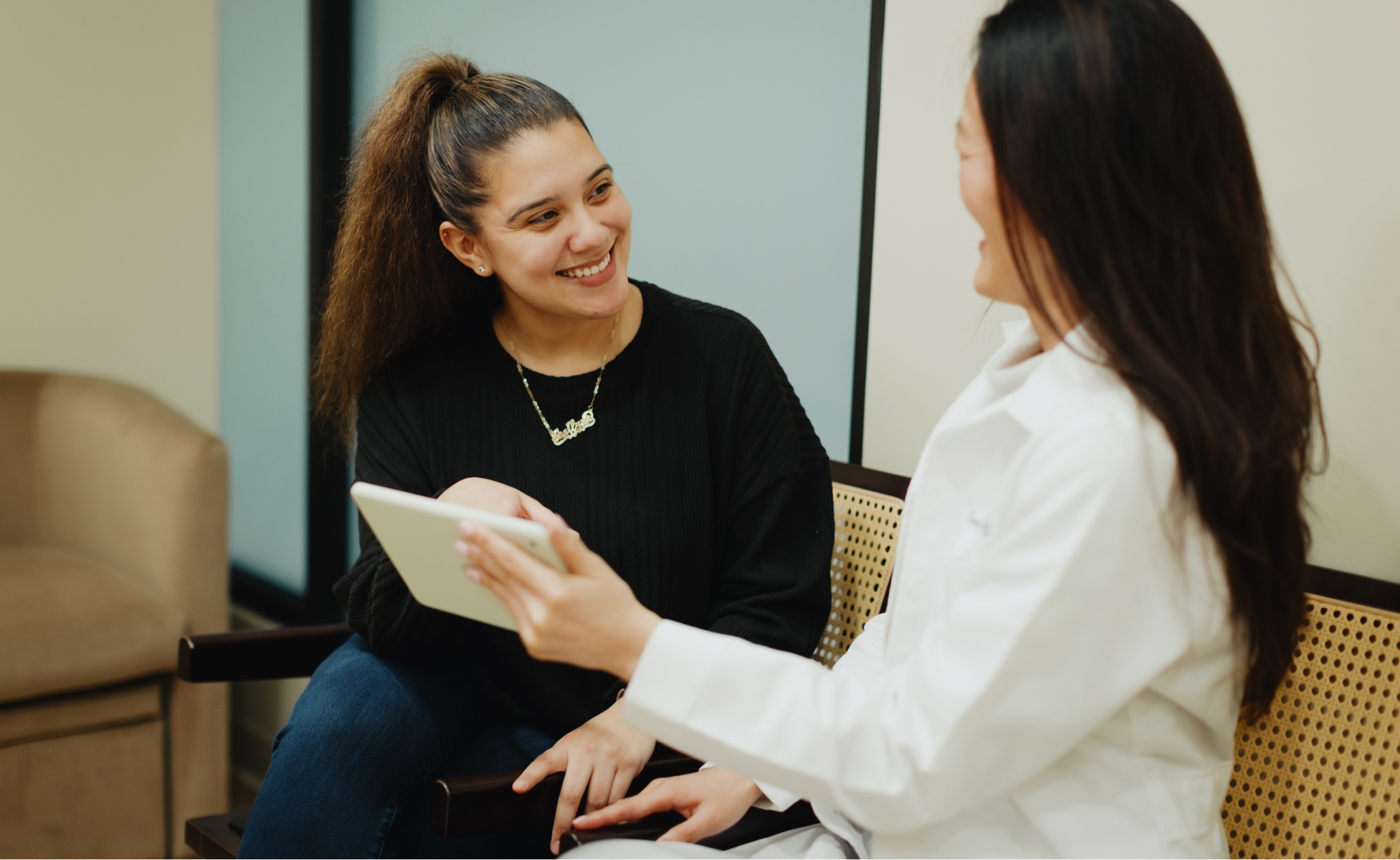 A woman in a white coat, possibly a dentist from Montvale Prosthodontics, holds a tablet and talks to a smiling woman with a ponytail seated on a bench in the waiting area.