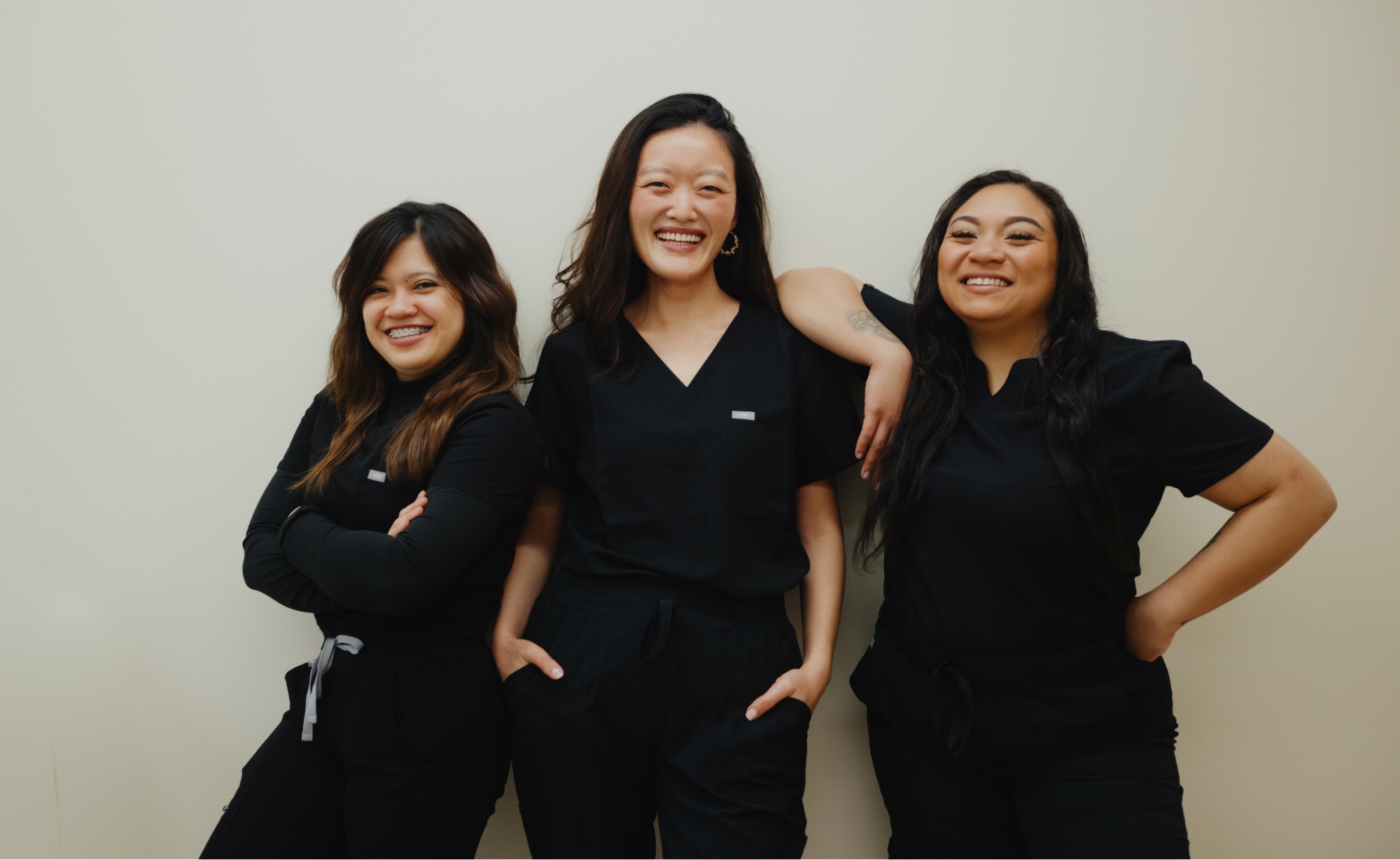 Three smiling women wearing black scrubs stand against a plain light-colored background, resembling a professional dental team. Two have their arms crossed while the third has her hands in her pockets.