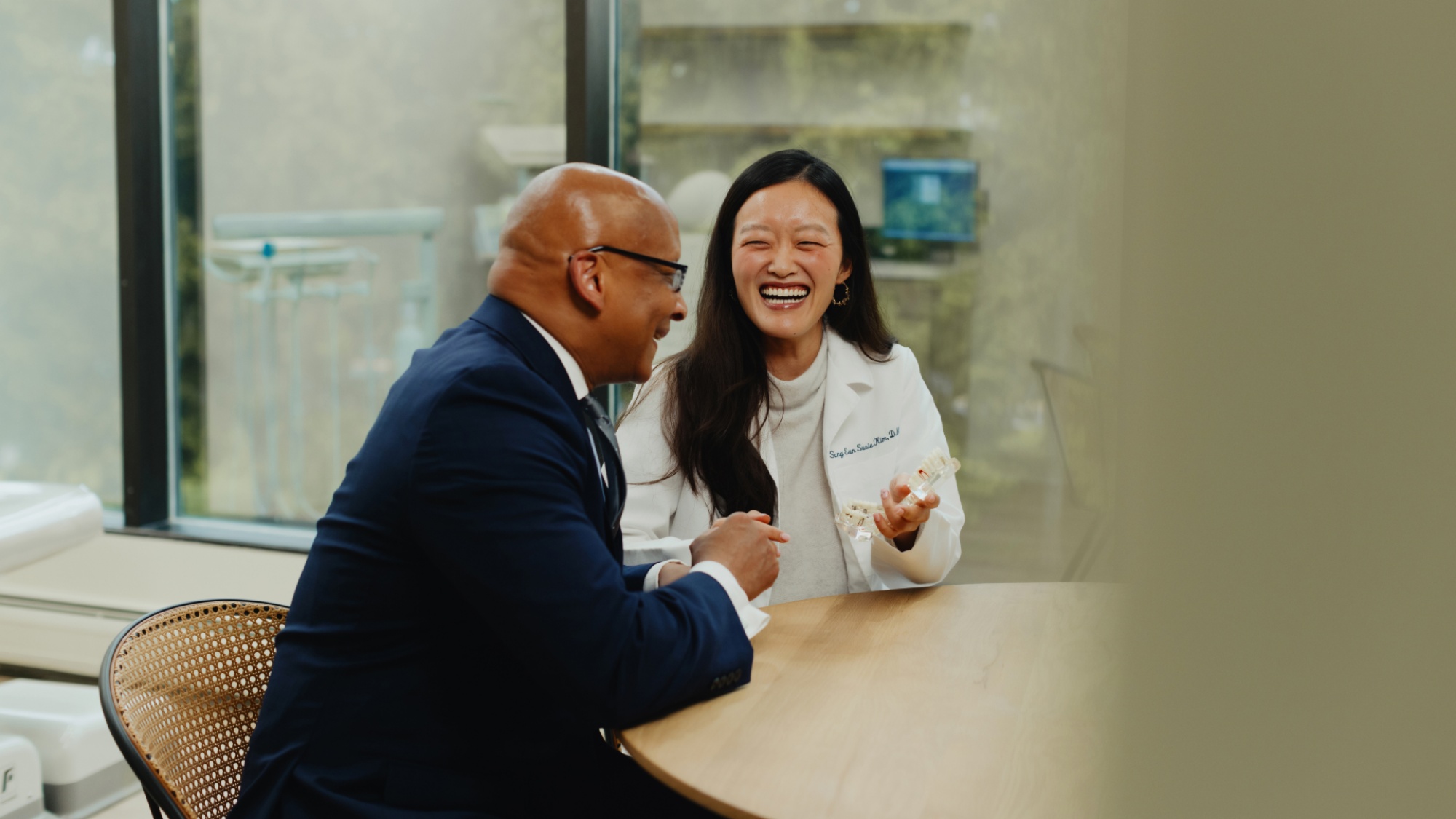 Two people are seated at a round table, engaged in conversation. The person on the left wears a suit, while the one on the right, a dentist from Montvale Prosthodontics wearing a lab coat, is smiling and holding a small object.