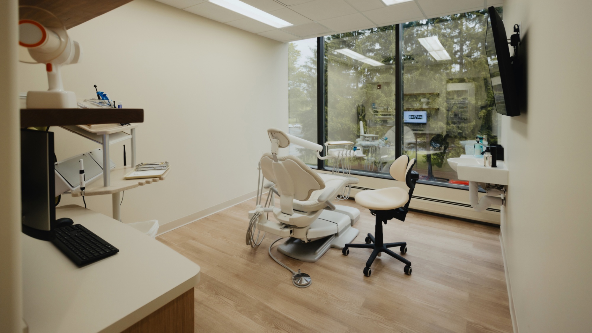 A modern dental examination room featuring a dentist's chair, instruments, a computer workstation, and a large window with a view of greenery outside.