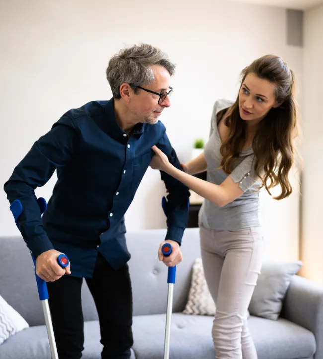 Une femme accompagne un homme en situation de convalescence à l’aide de béquilles. Monka soutient les aidants dans la gestion des soins et de l'autonomie à domicile.