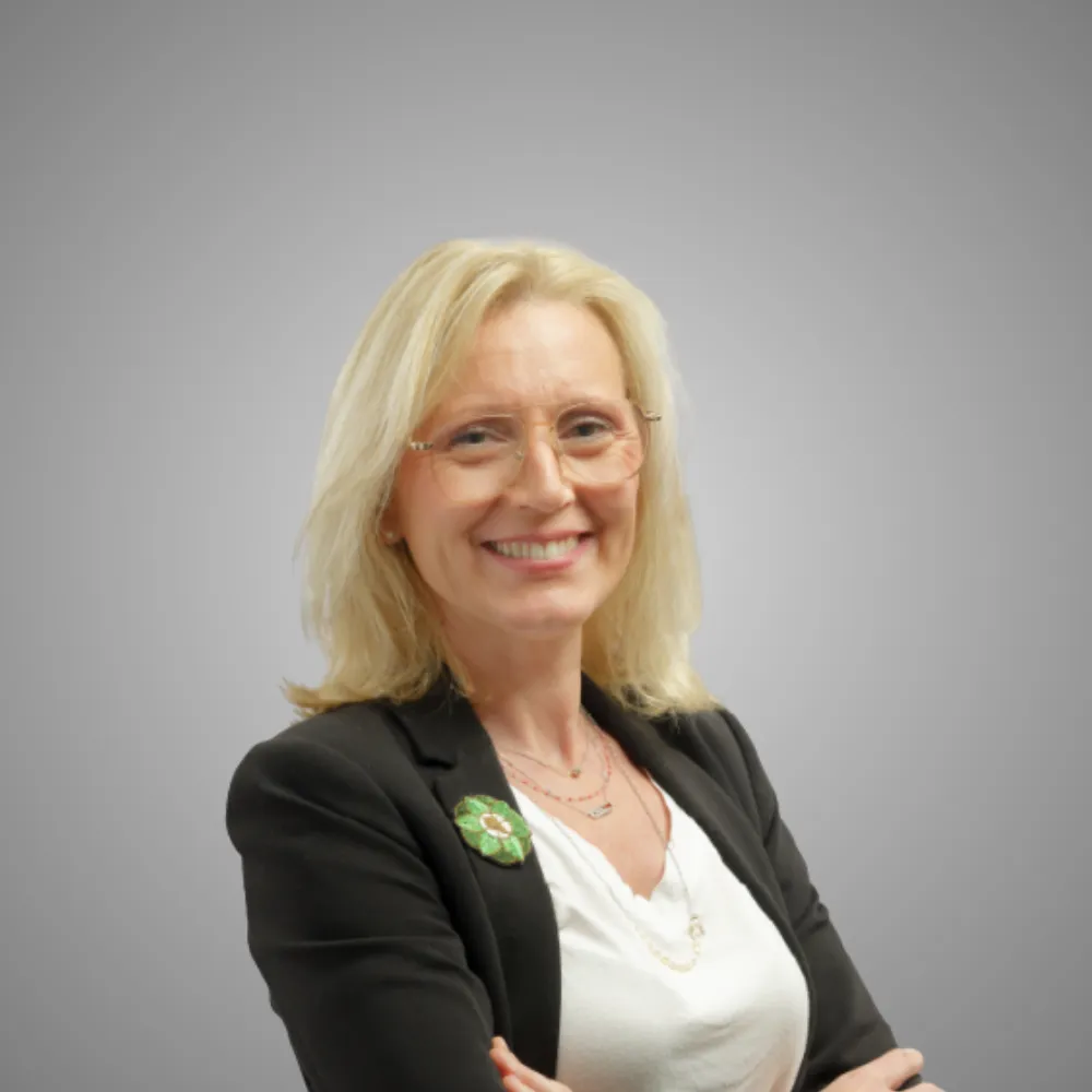 Femme souriante aux cheveux blond portant des lunettes, veste noire et blouse blanche.